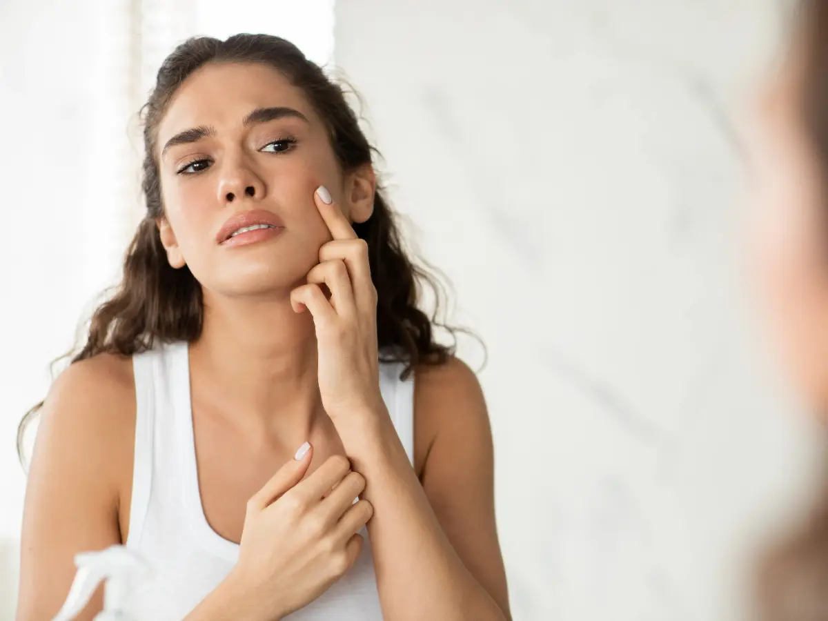  Frau schaut skeptisch in den Spiegel, betrachtet ihre Haut 