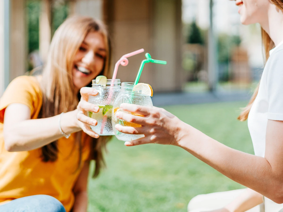  Zwei Frauen sitzen auf der Wiese und stoßen mit Getränk an 
