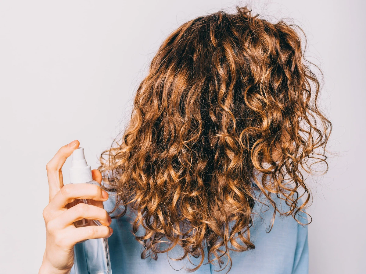  Junge Frau mit langen lockigen Haaren, die kosmetische Pflegeprodukte aufträgt. Spray mit Meersalz, um Beachy Waves Frisur zu machen. 