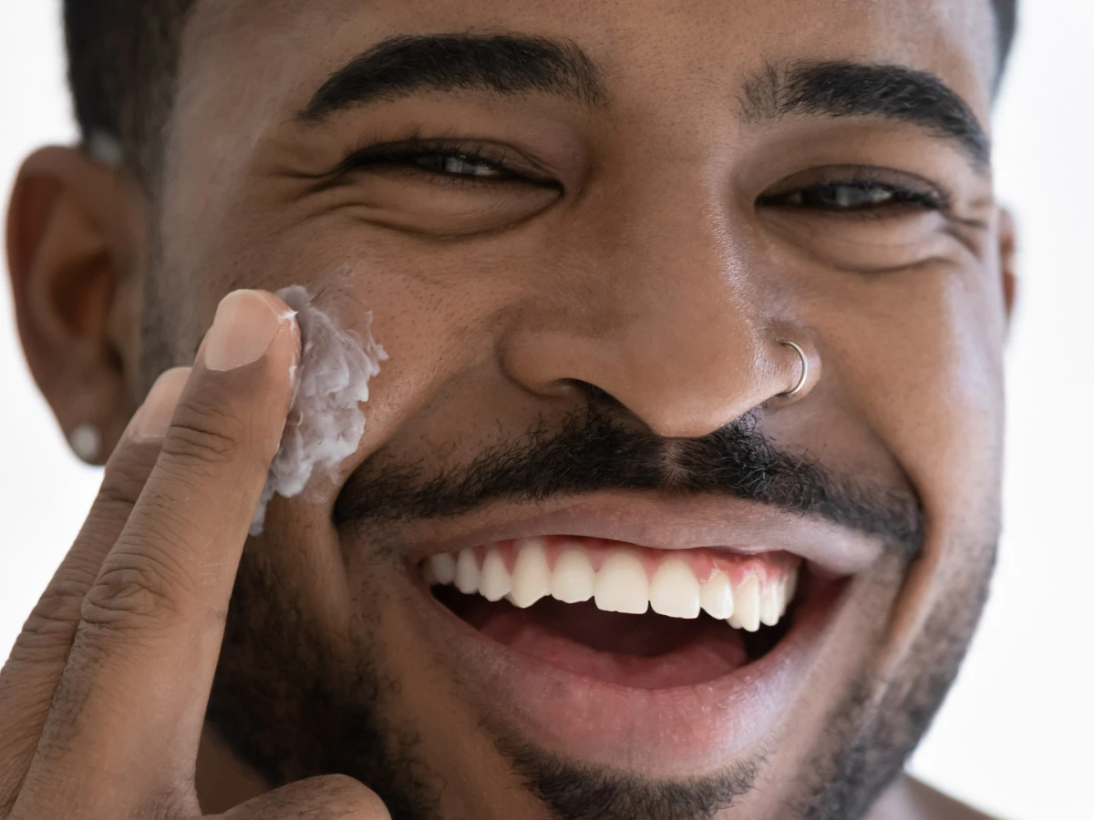  Junger Mann trägt Gesichtspflege auf und lacht 