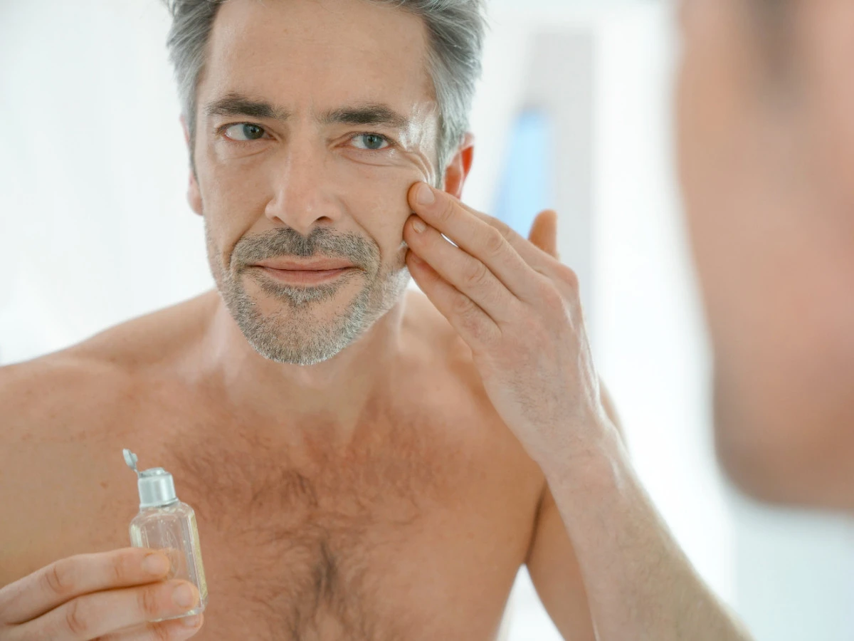  Älterer, reifer Mann steht vorm Spiegel und trägt Gesichtscreme auf 