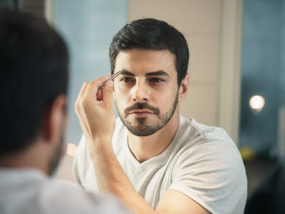  Man steht vor Spiegel und zupft sich die Augenbrauen 